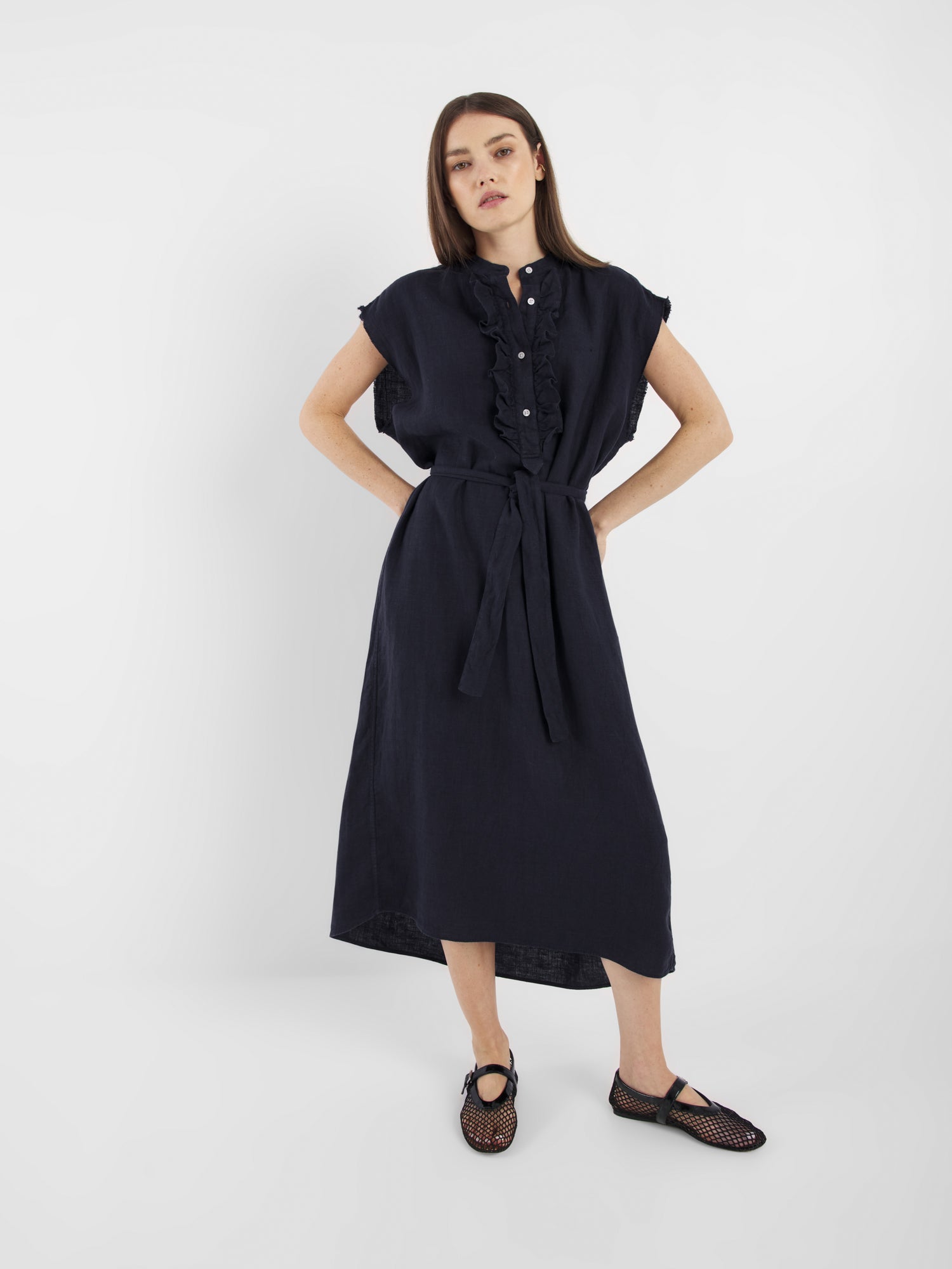 Eine Frau mit glattem braunem Haar und neutralem Gesichtsausdruck trägt das Dress Barcelona Navy - ein kurzärmeliges, geknöpftes, gegürtetes Midikleid mit Rüschendetails - in Kombination mit schwarzen Webschuhen vor einem schlichten weißen Hintergrund.