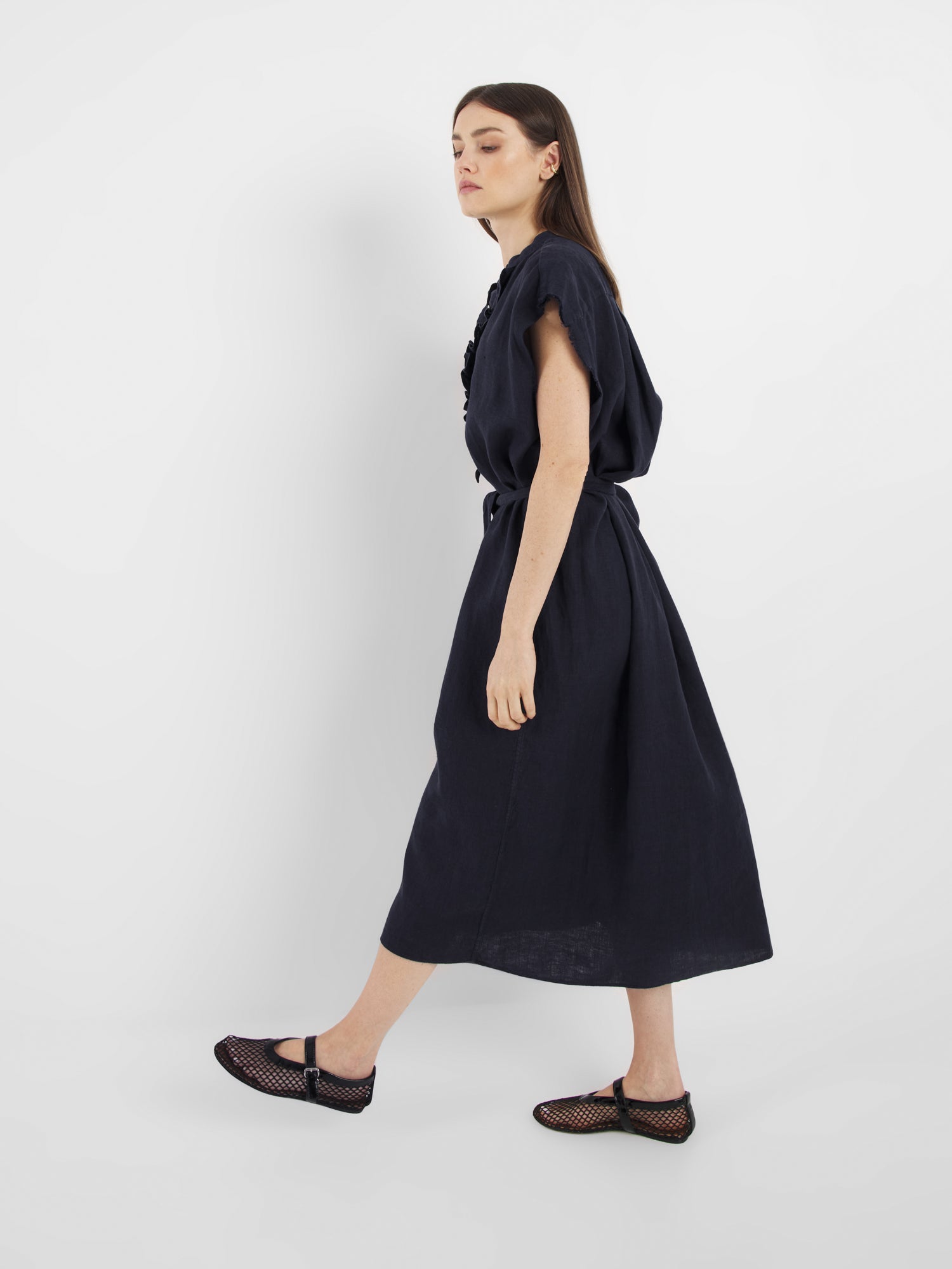 Eine Frau im Profil trägt das Dress Barcelona Navy, ein lockeres, kurzärmeliges Midikleid, gepaart mit schwarzen Netzschuhen vor einem schlichten weißen Hintergrund.