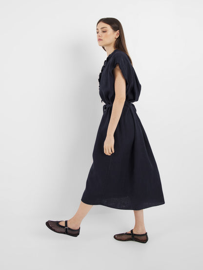 Eine Frau im Profil trägt das Dress Barcelona Navy, ein lockeres, kurzärmeliges Midikleid, gepaart mit schwarzen Netzschuhen vor einem schlichten weißen Hintergrund.