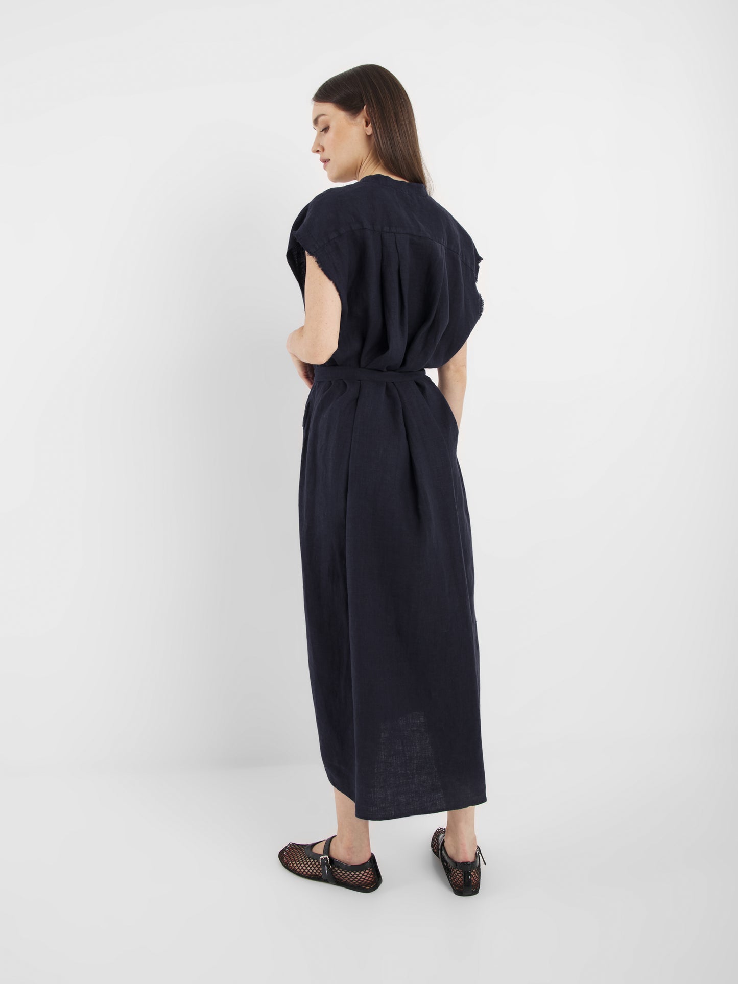 Eine Frau steht in einem minimalistischen weißen Raum und trägt das locker sitzende, kurzärmelige Kleid Barcelona Navy mit einer Taillenbinde. Sie blickt über die Schulter und kombiniert es mit schwarzen Mesh-Flats.