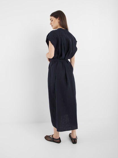 Eine Frau steht in einem minimalistischen weißen Raum und trägt das locker sitzende, kurzärmelige Kleid Barcelona Navy mit einer Taillenbinde. Sie blickt über die Schulter und kombiniert es mit schwarzen Mesh-Flats.