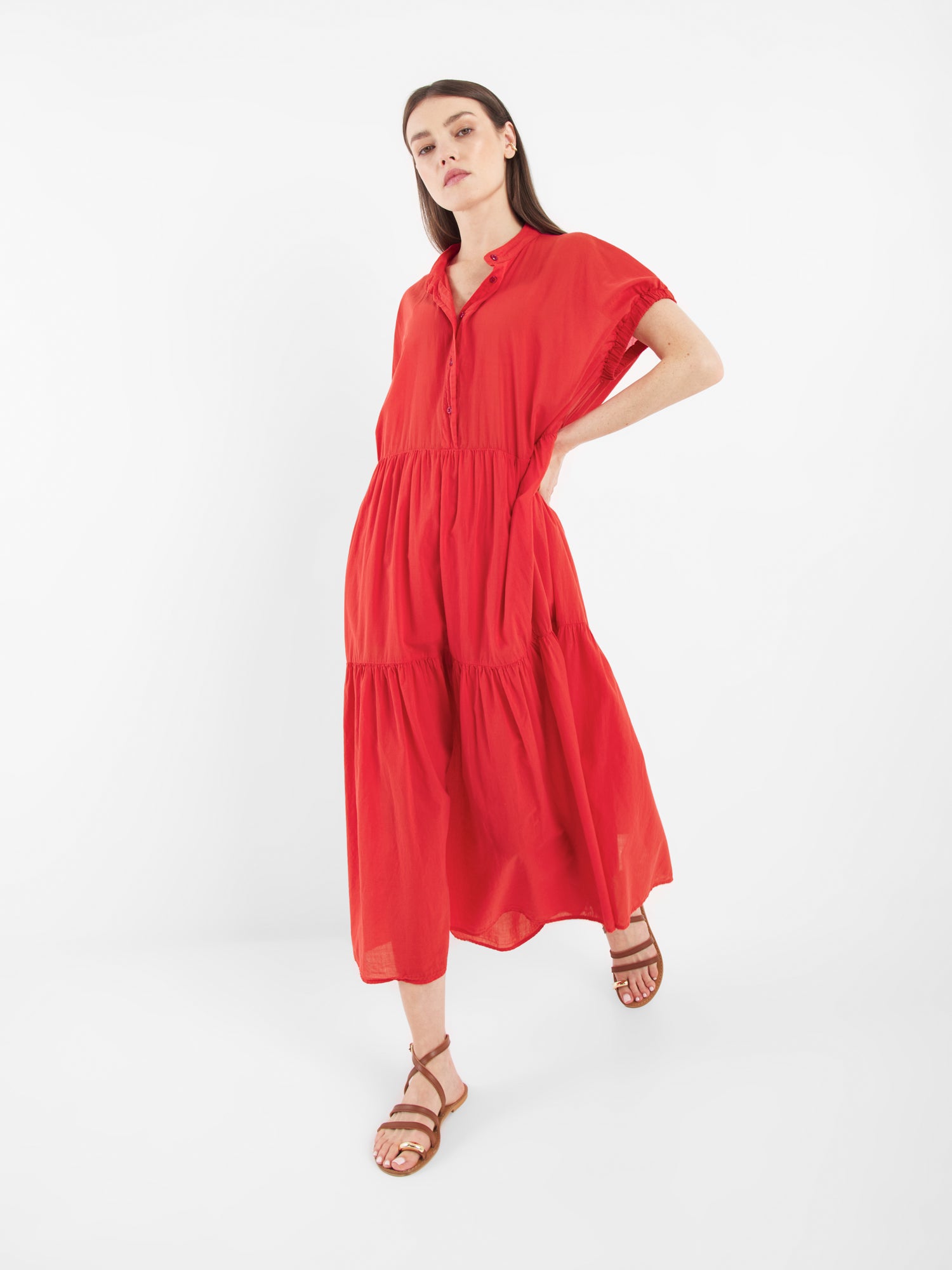 Eine Frau posiert entspannt mit dem Kleid Cowboy Boho Red und braunen Riemchensandalen vor einem schlichten weißen Hintergrund.