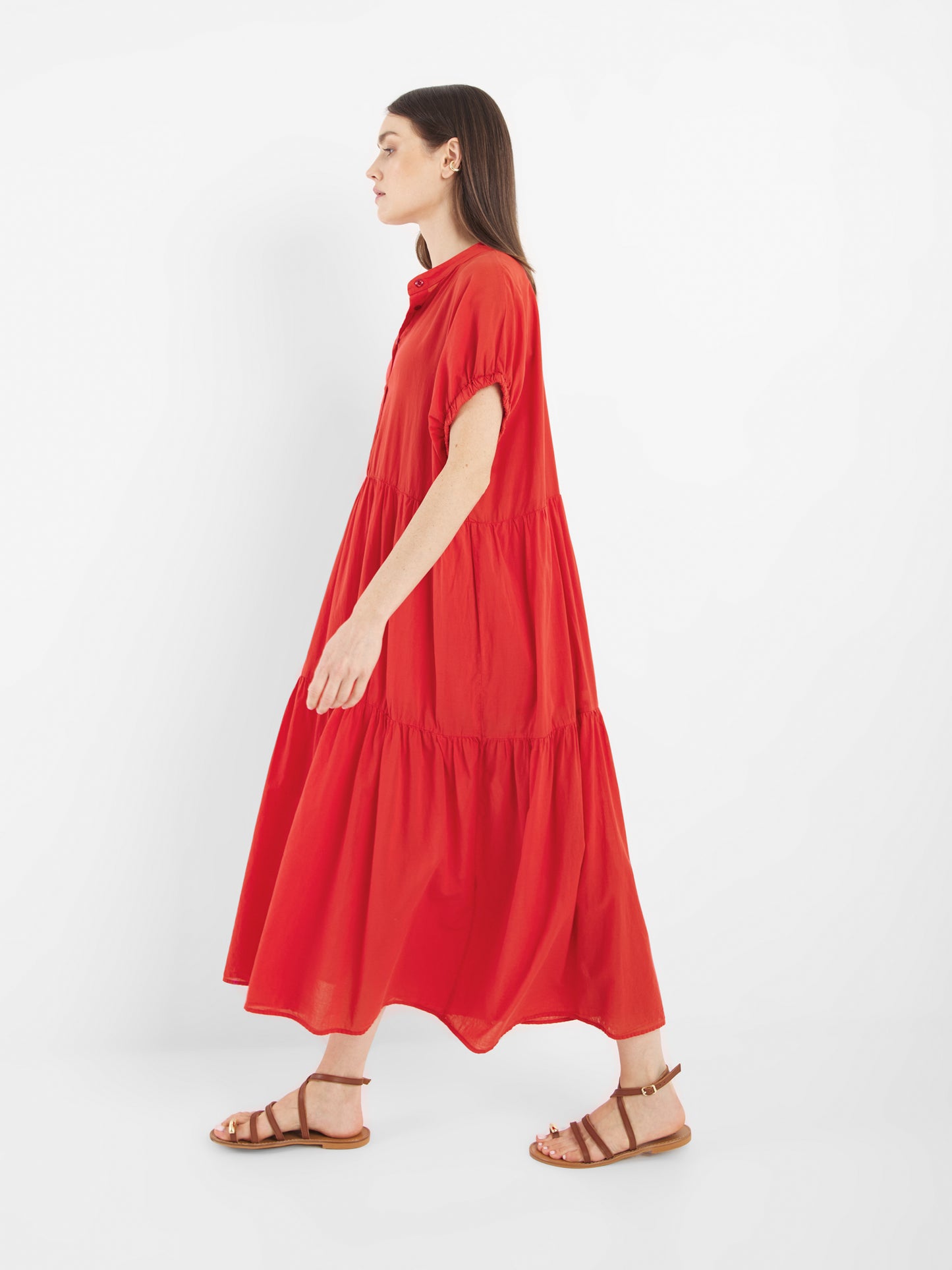 Frau im Profil, die das Kleid Cowboy Boho Red trägt, ein locker sitzendes, gestuftes Maxikleid mit kurzen Ärmeln und braunen flachen Sandalen, vor einem schlichten weißen Hintergrund.