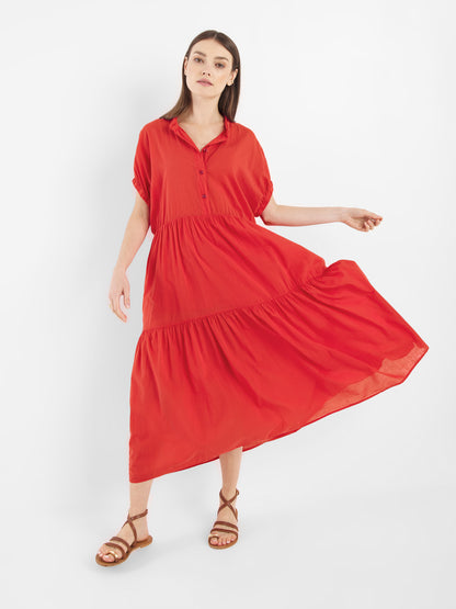 Eine Frau trägt das Kleid Cowboy Boho Red - ein lockeres, gestuftes Midikleid mit kurzen Ärmeln und einem Kragen - mit braunen Riemchensandalen, steht vor einem weißen Hintergrund und hält einen Teil ihres Kleides zur Seite.