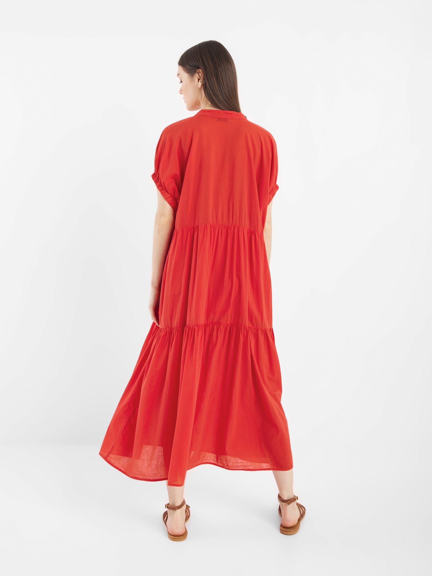 Eine Frau mit langen braunen Haaren, die das Dress Cowboy Boho Red trägt - ein knöchellanges, locker sitzendes, leuchtend rotes Kleid mit kurzen Ärmeln und gestuften Lagen - steht in braunen Sandalen vor einem schlichten weißen Hintergrund.