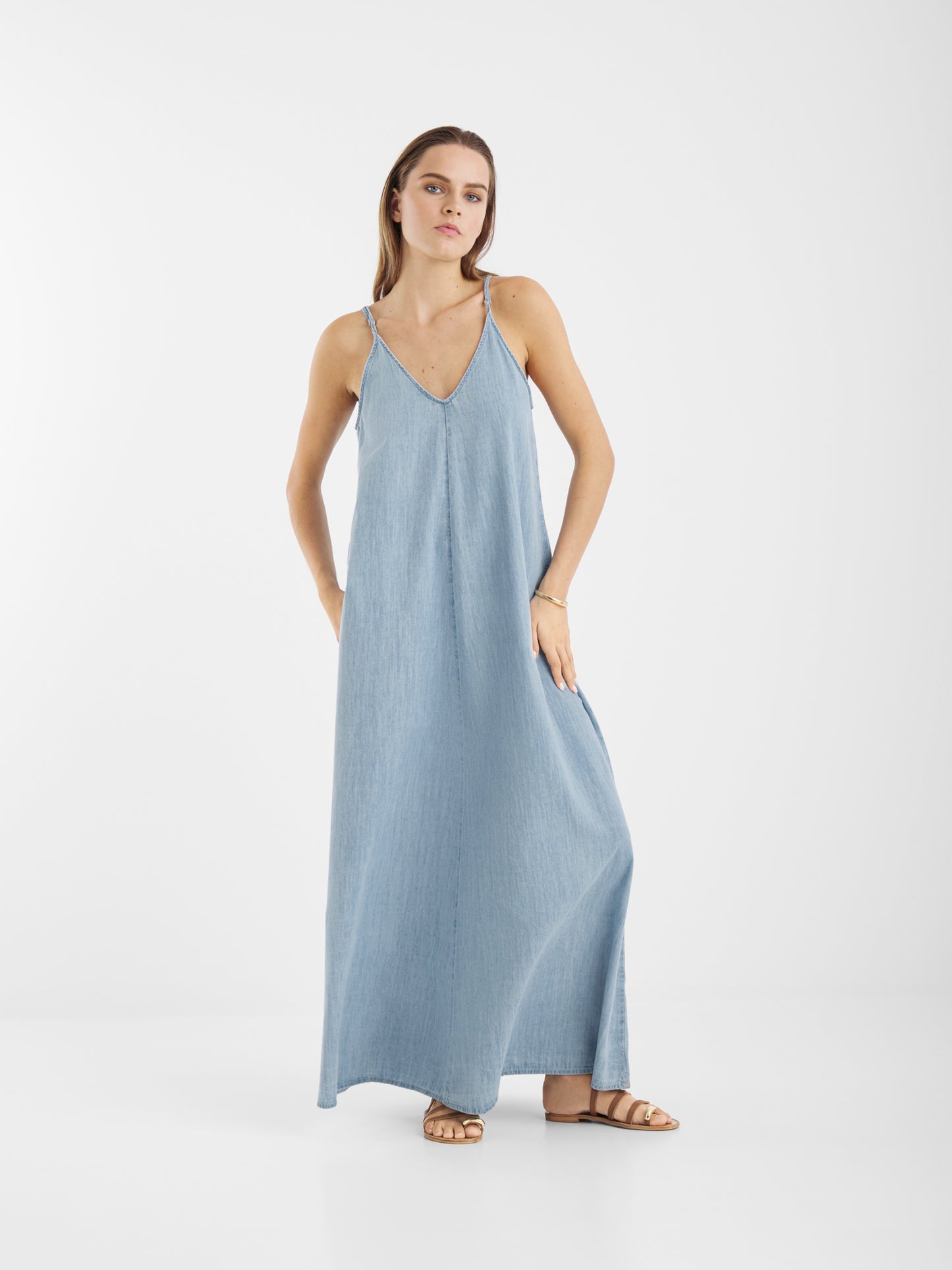 Eine Frau steht vor einem schlichten weißen Hintergrund und trägt das Dress Denim Tank, ein hellblaues, ärmelloses Maxikleid mit V-Ausschnitt, dünnen Trägern und Taschen. Sie kombiniert es mit flachen braunen Sandalen und hat die Hände in den Taschen des Kleides.
