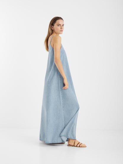 Eine Frau in einem hellblauen Dress Denim Tank und braunen Sandalen steht seitlich vor einem schlichten weißen Hintergrund und schaut in die Kamera.