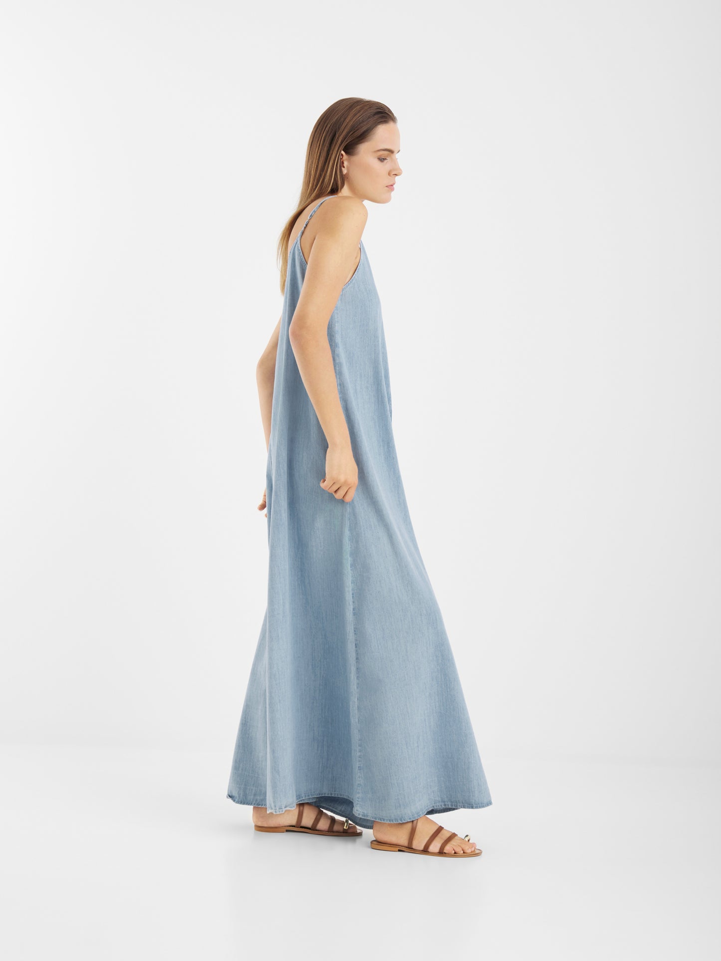Eine Frau modelliert das Dress Denim Tank, ein ärmelloses hellblaues Maxikleid, gepaart mit braunen flachen Sandalen, während sie vor einem schlichten weißen Hintergrund steht und ihren Körper leicht zur Seite neigt.