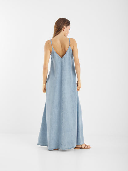Eine Frau mit langen braunen Haaren steht mit dem Blick zur Seite und trägt das Dress Denim Tank - ein ärmelloses, hellblaues, knöchellanges Jeanskleid mit dünnen Trägern und tiefem V-Ausschnitt -, gepaart mit flachen Sandalen, vor einem schlichten weißen Hintergrund.