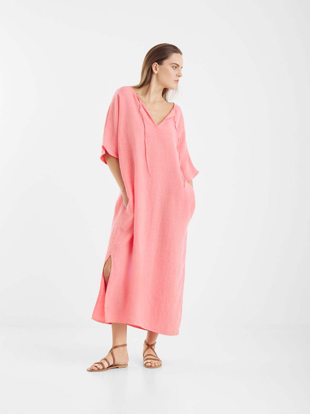 Eine Frau steht vor einem weißen Hintergrund und trägt das Let's Go Marrakesch Kaftan Kleid, einen knöchellangen, locker sitzenden Kaftan in Hellrosa mit Seitenschlitzen, Dreiviertelärmeln, Taschen und braunen flachen Sandalen.