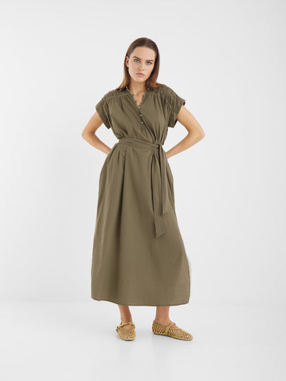 Eine Frau steht vor einem weißen Hintergrund und trägt das Kleid Maui - ein olivgrünes, kurzärmeliges Maxikleid aus Baumwolle. Sie kombiniert es mit gewebten braunen Halbschuhen. Ihr glattes braunes Haar und ihr neutraler Gesichtsausdruck vervollständigen den Look.