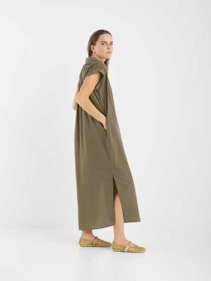 Eine Frau mit hellbraunem Haar steht seitlich und trägt ein lockeres, olivgrünes Kleid Maui mit kurzen Ärmeln und Seitenschlitzen, gepaart mit gewebten beigen Flats vor einem schlichten weißen Hintergrund.