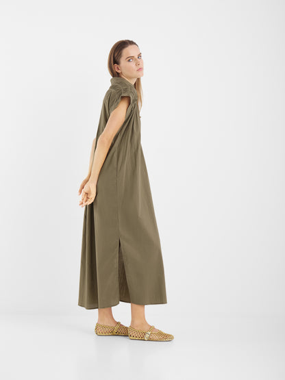 Eine Frau in einem olivgrünen Dress Maui, einem kurzärmeligen Maxikleid aus Portugal, steht seitlich mit den Händen auf dem Rücken, trägt hellbraune gewebte Slipper und blickt nach oben auf einen schlichten weißen Hintergrund.