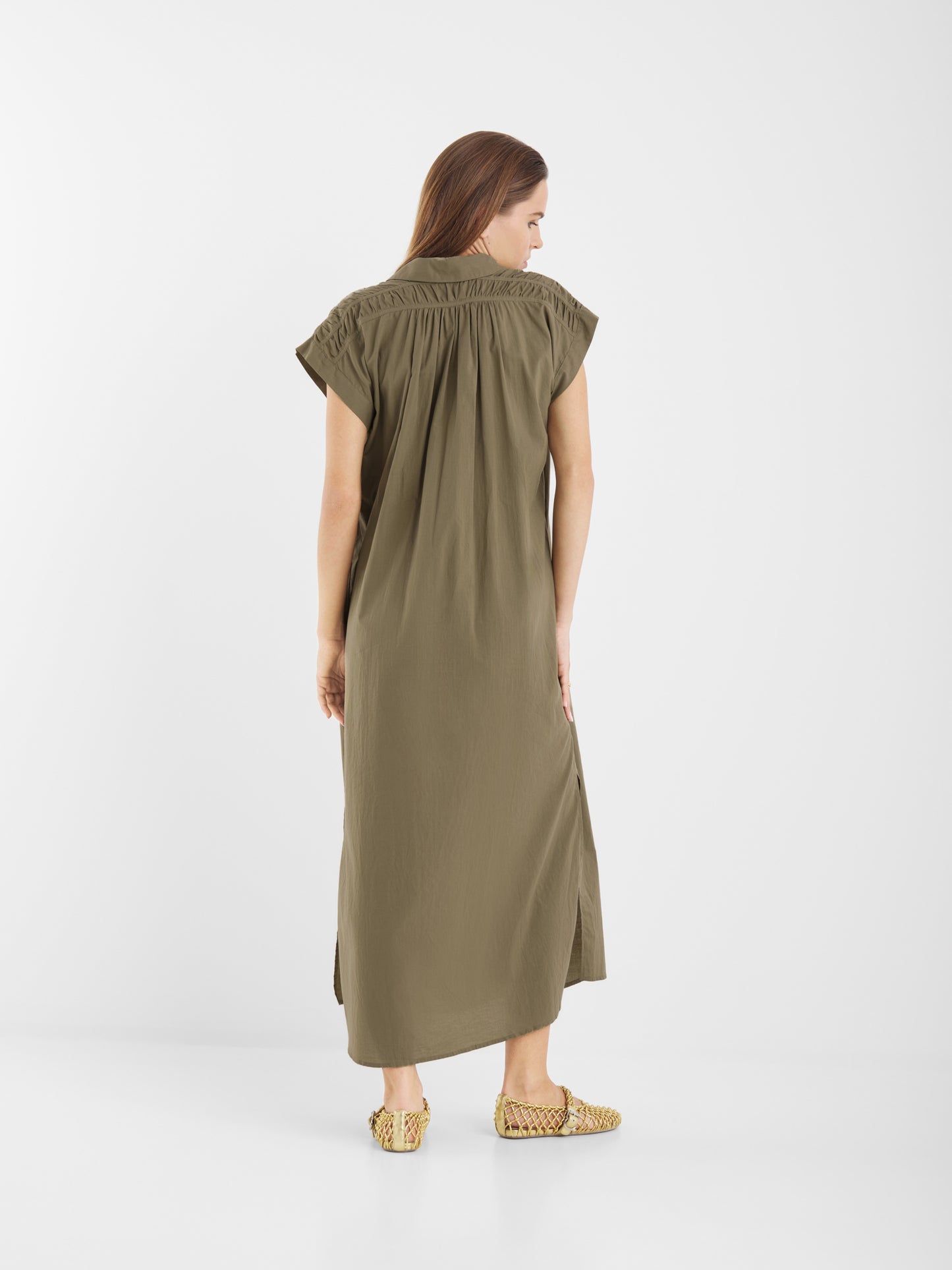 Eine Frau blickt von sich weg, sie trägt ein locker sitzendes olivgrünes Kleid Maui mit kurzen Ärmeln und gewebten hellbraunen Slippern vor einem schlichten weißen Hintergrund.