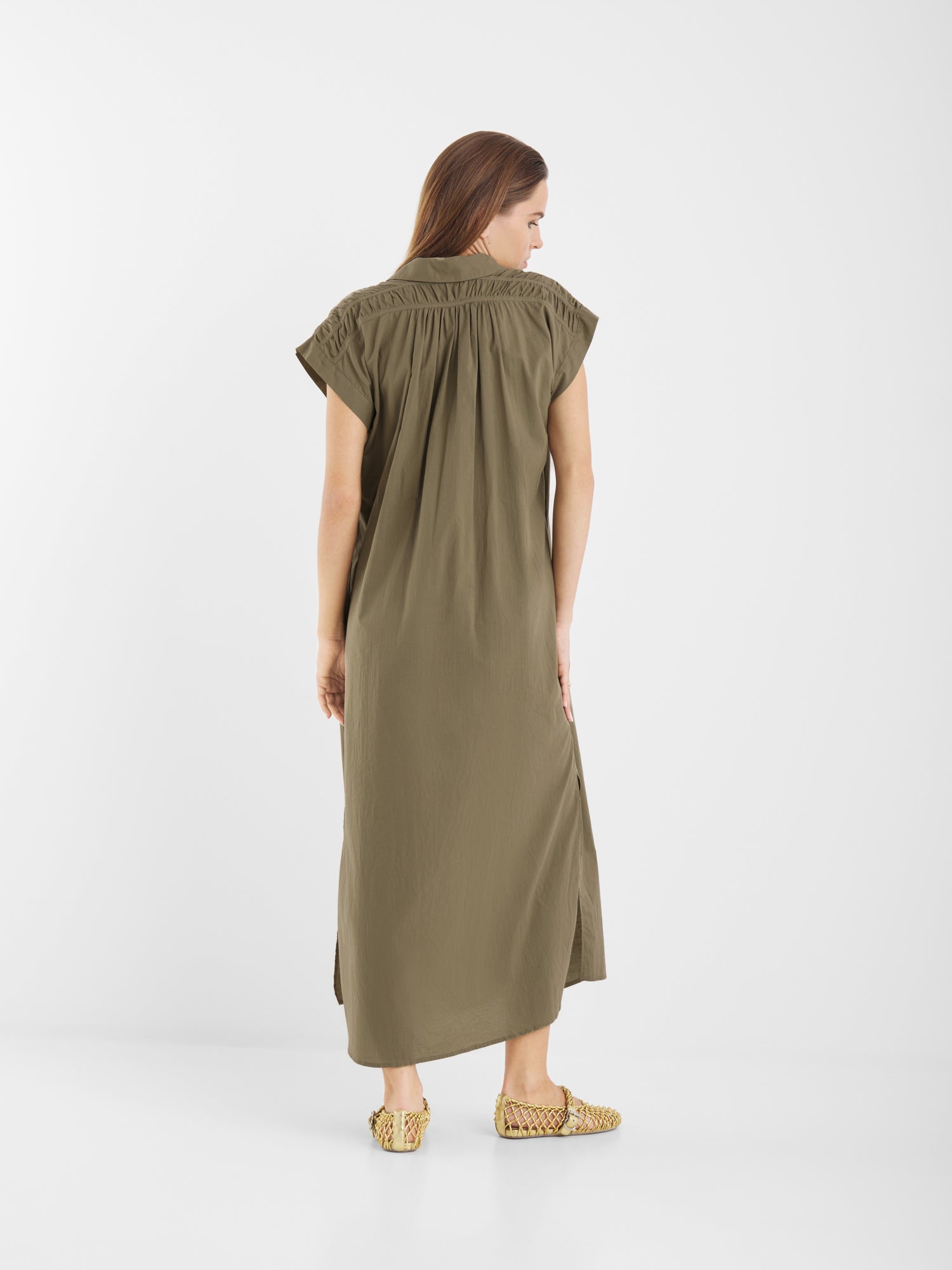 Eine Frau blickt von sich weg, sie trägt ein locker sitzendes olivgrünes Kleid Maui mit kurzen Ärmeln und gewebten hellbraunen Slippern vor einem schlichten weißen Hintergrund.