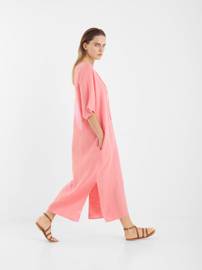 Eine Frau mit glattem hellbraunem Haar und ernster Miene geht in hellbraunen Sandalen seitwärts und trägt das Kleid Kaftan Linen Pink - ein lockeres Leinenkleid mit kurzen Ärmeln und Seitenschlitzen - vor einem schlichten weißen Hintergrund.