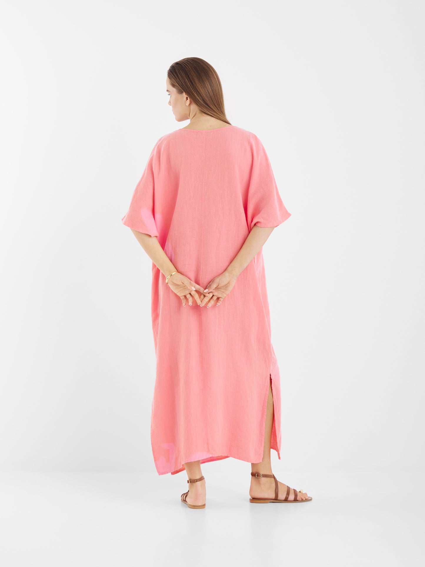 Eine Frau mit langen braunen Haaren steht mit dem Rücken zur Wand und trägt das nachhaltig produzierte Kleid Kaftan Leinen Pink mit kurzen Ärmeln und Seitenschlitzen. Sie trägt flache braune Sandalen, die Hände hinter dem Rücken, vor einem weißen Hintergrund.