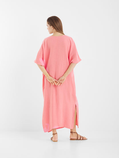 Eine Frau mit langen braunen Haaren steht mit dem Rücken zur Wand und trägt das nachhaltig produzierte Kleid Kaftan Leinen Pink mit kurzen Ärmeln und Seitenschlitzen. Sie trägt flache braune Sandalen, die Hände hinter dem Rücken, vor einem weißen Hintergrund.