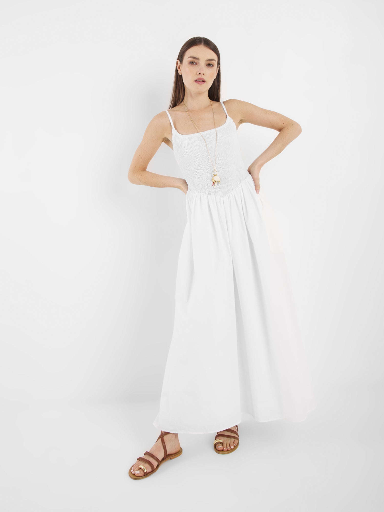 Eine Frau mit glattem braunem Haar und neutralem Gesichtsausdruck steht vor einem weißen Hintergrund, die Hände in die Hüften gestemmt, und trägt das Elisa Dress White, braune Sandalen und eine lange Halskette mit Anhänger.