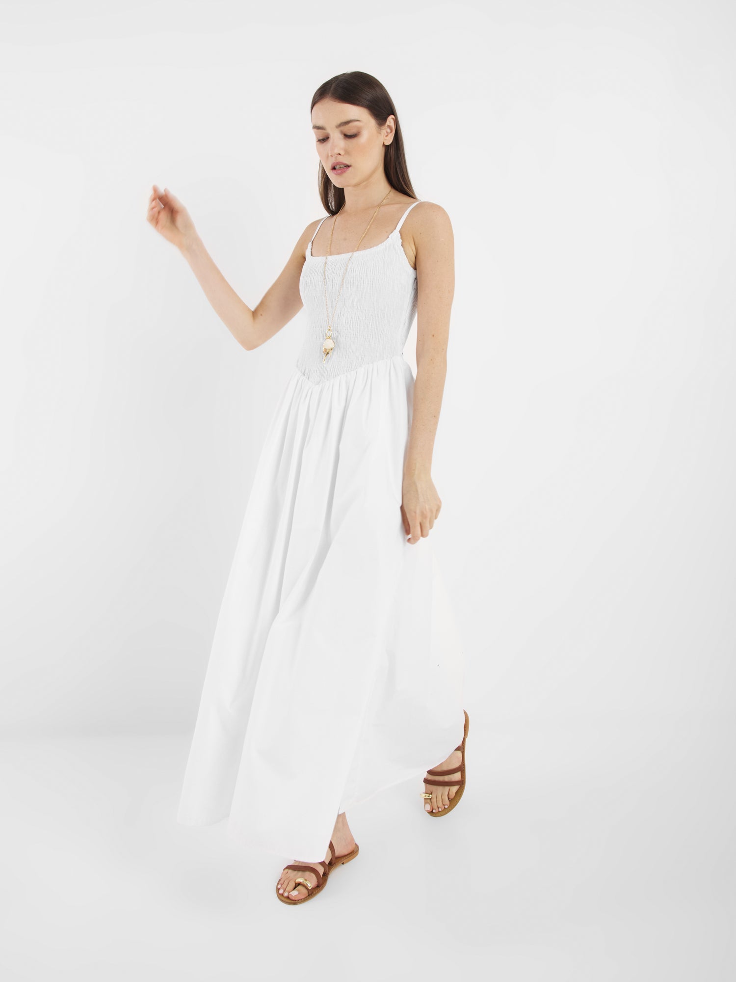 Frau trägt das Elisa Dress White, ein langes, ärmelloses Kleid mit Spaghettiträgern, kombiniert mit braunen Sandalen und einer langen Halskette, vor einem schlichten weißen Hintergrund.