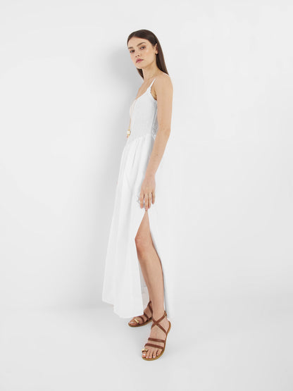 Eine Frau steht im Profil vor einem schlichten weißen Hintergrund. Sie trägt das Elisa Dress White - ein ärmelloses Kleid mit hohem Schlitz - und dazu braune Riemchensandalen.