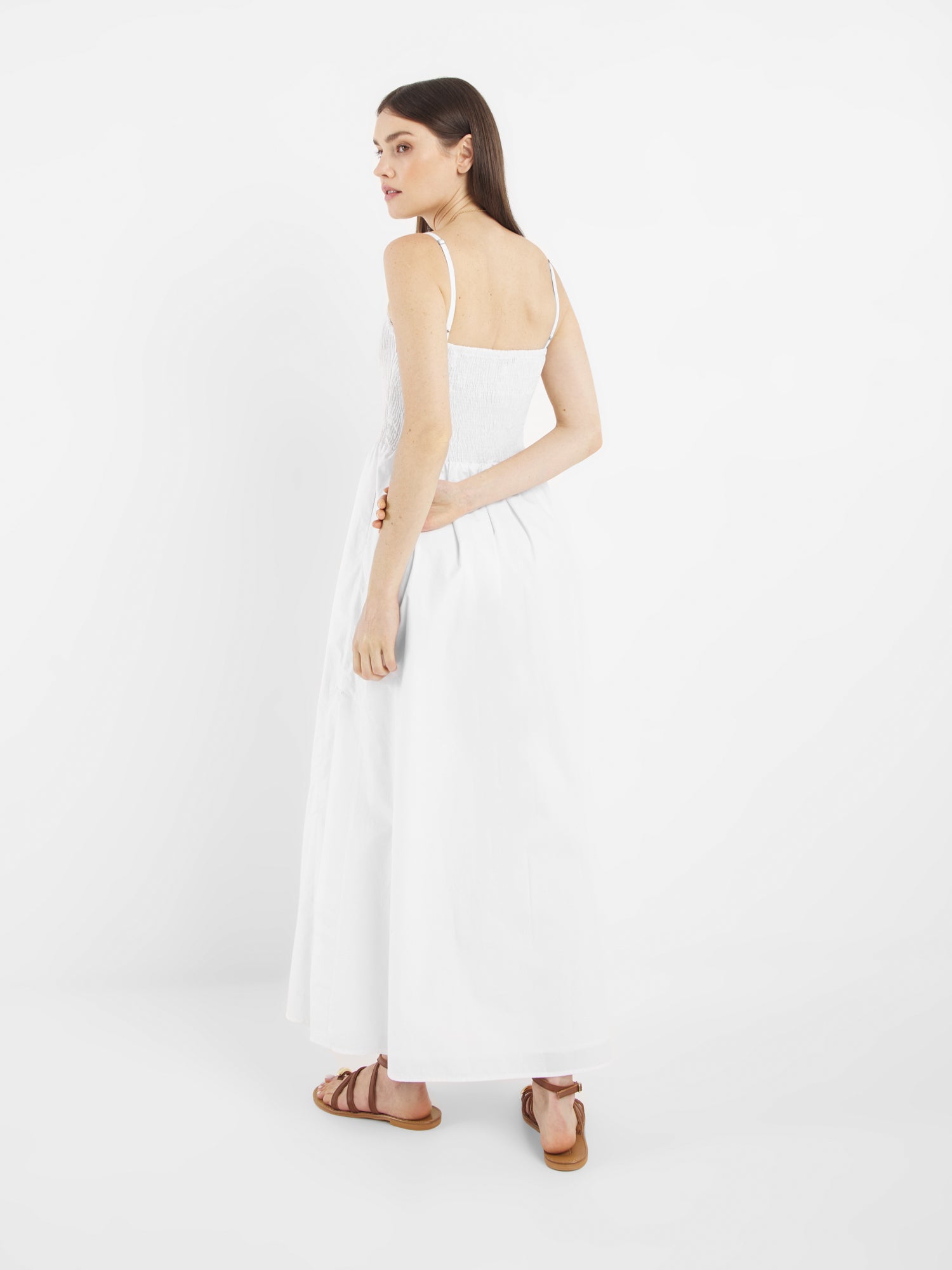 Eine Frau mit langen braunen Haaren trägt das Elisa Dress White - ein knöchellanges, ärmelloses weißes Kleid - mit braunen Sandalen, steht vor einem weißen Hintergrund und blickt über ihre Schulter.