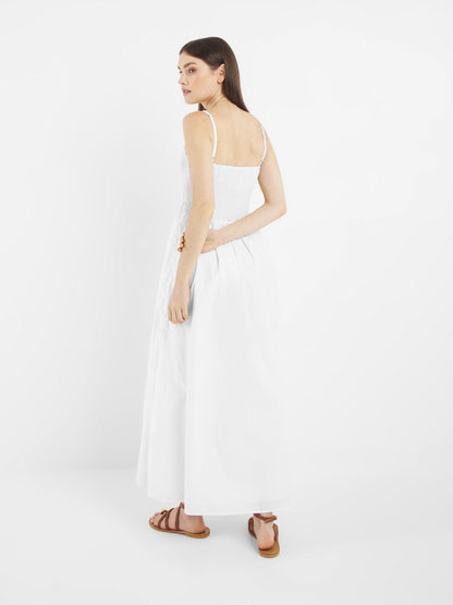 Eine Frau mit langen braunen Haaren trägt das Elisa Dress White - ein knöchellanges, ärmelloses weißes Kleid - mit braunen Sandalen, steht vor einem weißen Hintergrund und blickt über ihre Schulter.