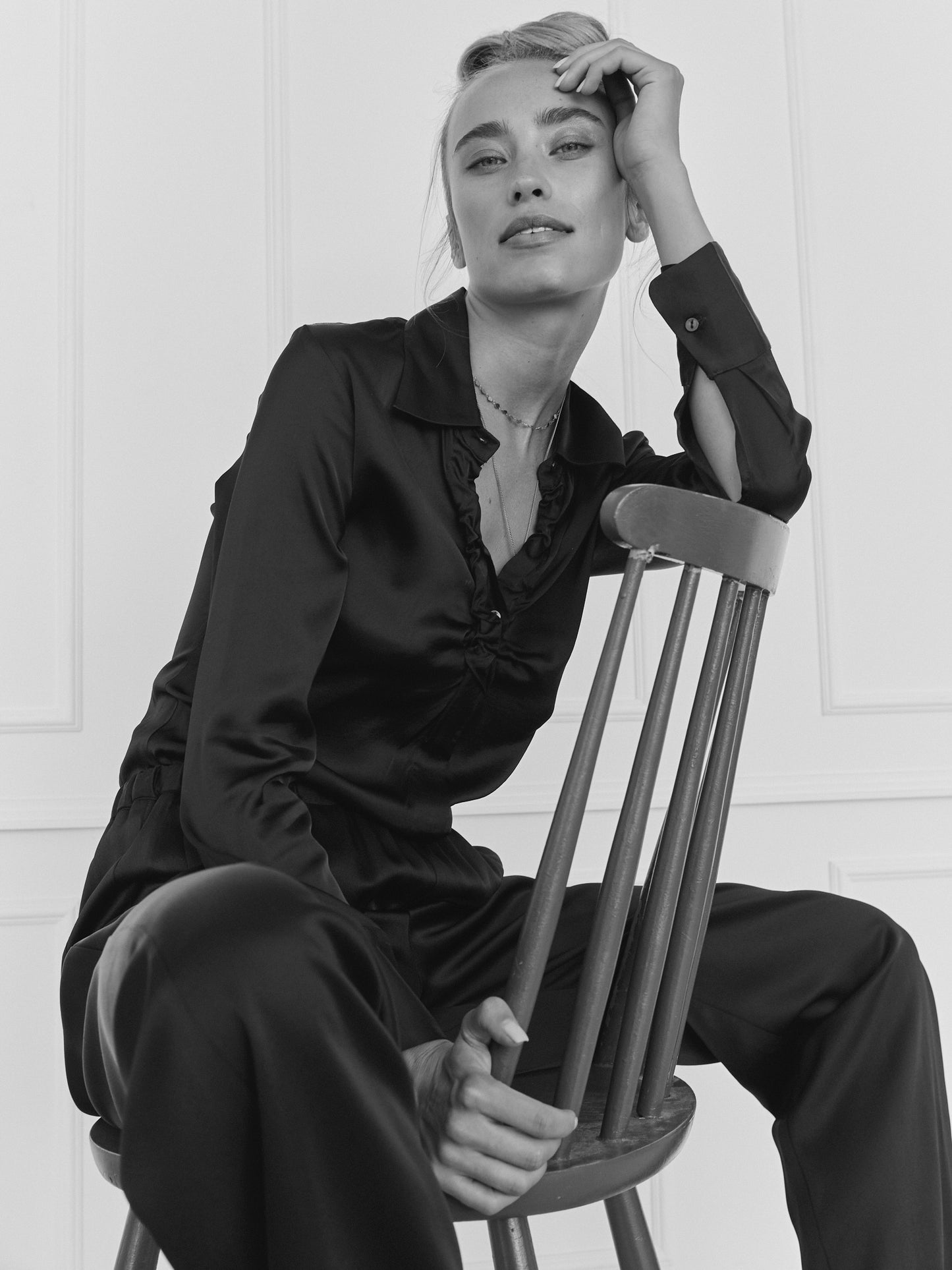Eine Frau, die die Bluse Ella Shiny Black und die dazu passende Hose trägt, sitzt auf einem Holzstuhl und blickt sanft in die Kamera, während sie ihre Hand auf die Stirn legt. Das schwarz-weiße Foto hat einen minimalistischen Hintergrund.