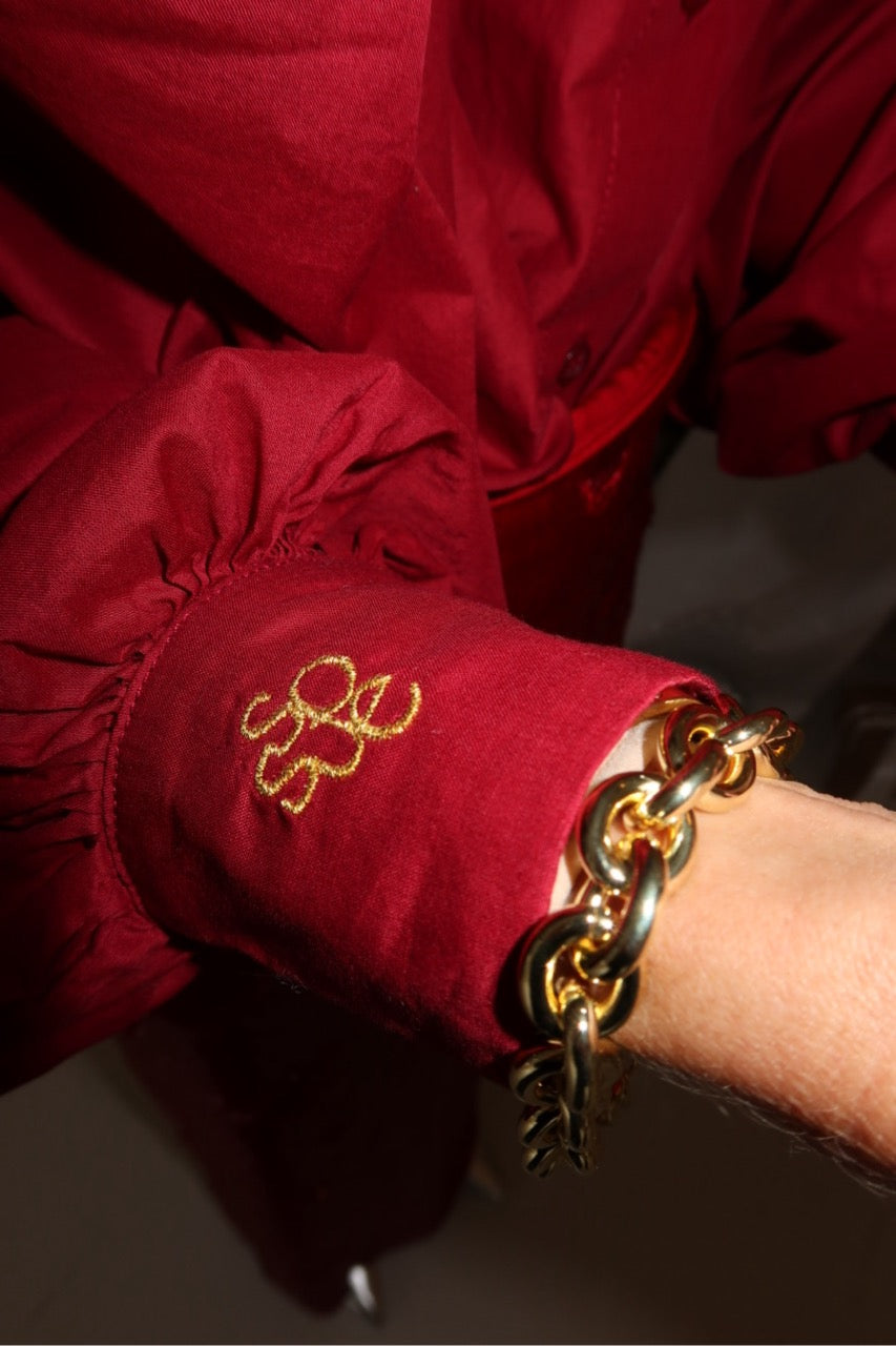 Eine Person trägt die Bluse Antonia Burgundy - eine burgunderfarbene Damenbluse mit gelber Manschettenstickerei - in Kombination mit einem klobigen gold-schwarzen Kettenarmband.