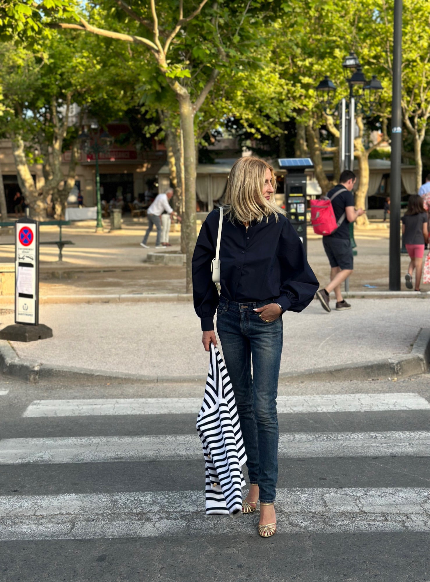 Eine blonde Frau steht auf einem Zebrastreifen und trägt ein gestreiftes Hemd. Sie trägt die Bluse Antonia Slim Dark Blue, Jeans und goldene Schuhe. Bäume, Menschen und Geschäfte sind an einem sonnigen Tag im Hintergrund zu sehen.