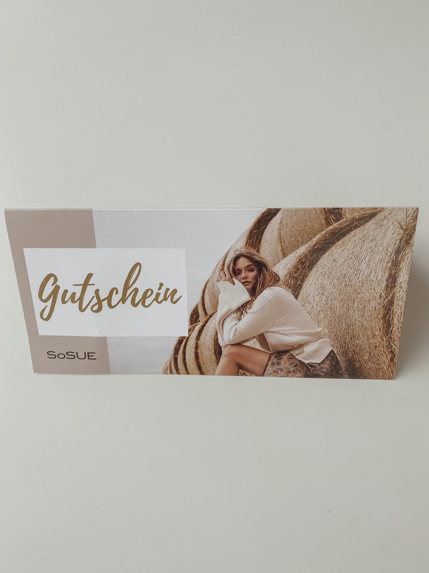 Eine Geschenkgutschein-Karte mit dem Wort "Gutschein" und einer Frau in einem hellen Pullover auf Heuballen vor weißem Hintergrund - eine stilvolle Mode-Geschenkidee.