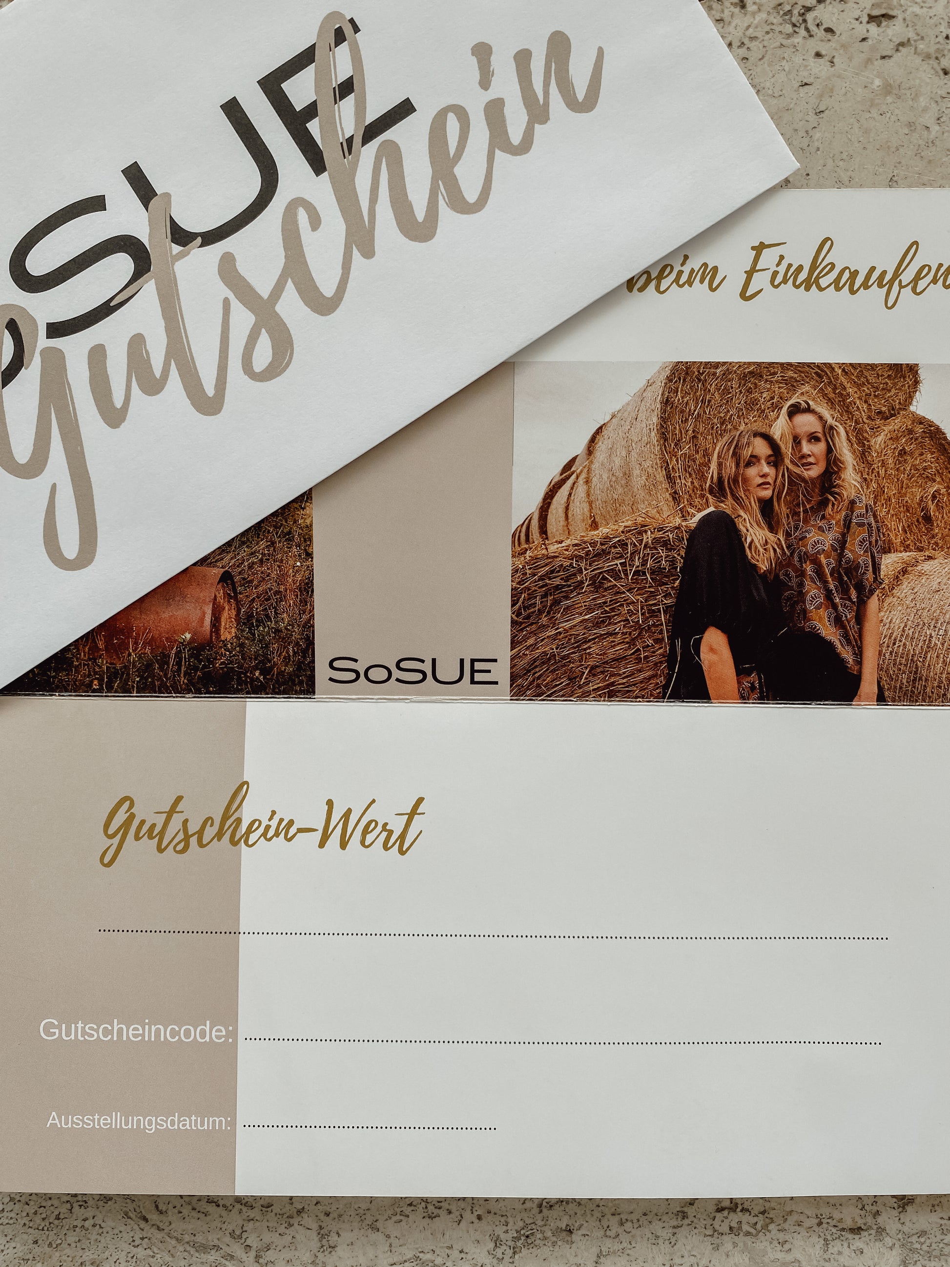 Ein Geschenkgutschein auf einer Steinoberfläche mit einem Umschlag und einer Karte, die zwei Frauen neben Heuballen zeigt, mit Feldern für Gutscheinwert, Code und Datum. Die perfekte Mode-Geschenkidee!.