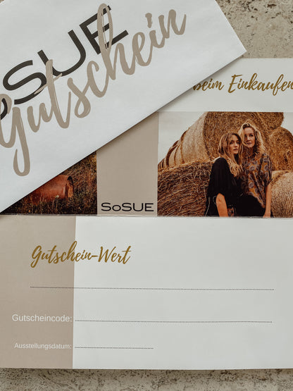 Ein Geschenkgutschein auf einer Steinoberfläche mit einem Umschlag und einer Karte, die zwei Frauen neben Heuballen zeigt, mit Feldern für Gutscheinwert, Code und Datum. Die perfekte Mode-Geschenkidee!.