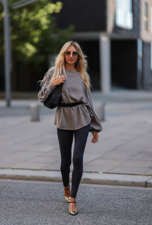 Eine Frau mit langen blonden Haaren geht mit einer Sonnenbrille, einem lockeren grauen Pullover mit Gürtel in der Taille, einer Hose Leggings Grey-80% Recycled und gemusterten Absätzen nach draußen und trägt eine schwarze Tasche. Städtische Szene mit Bäumen und modernen Gebäuden.