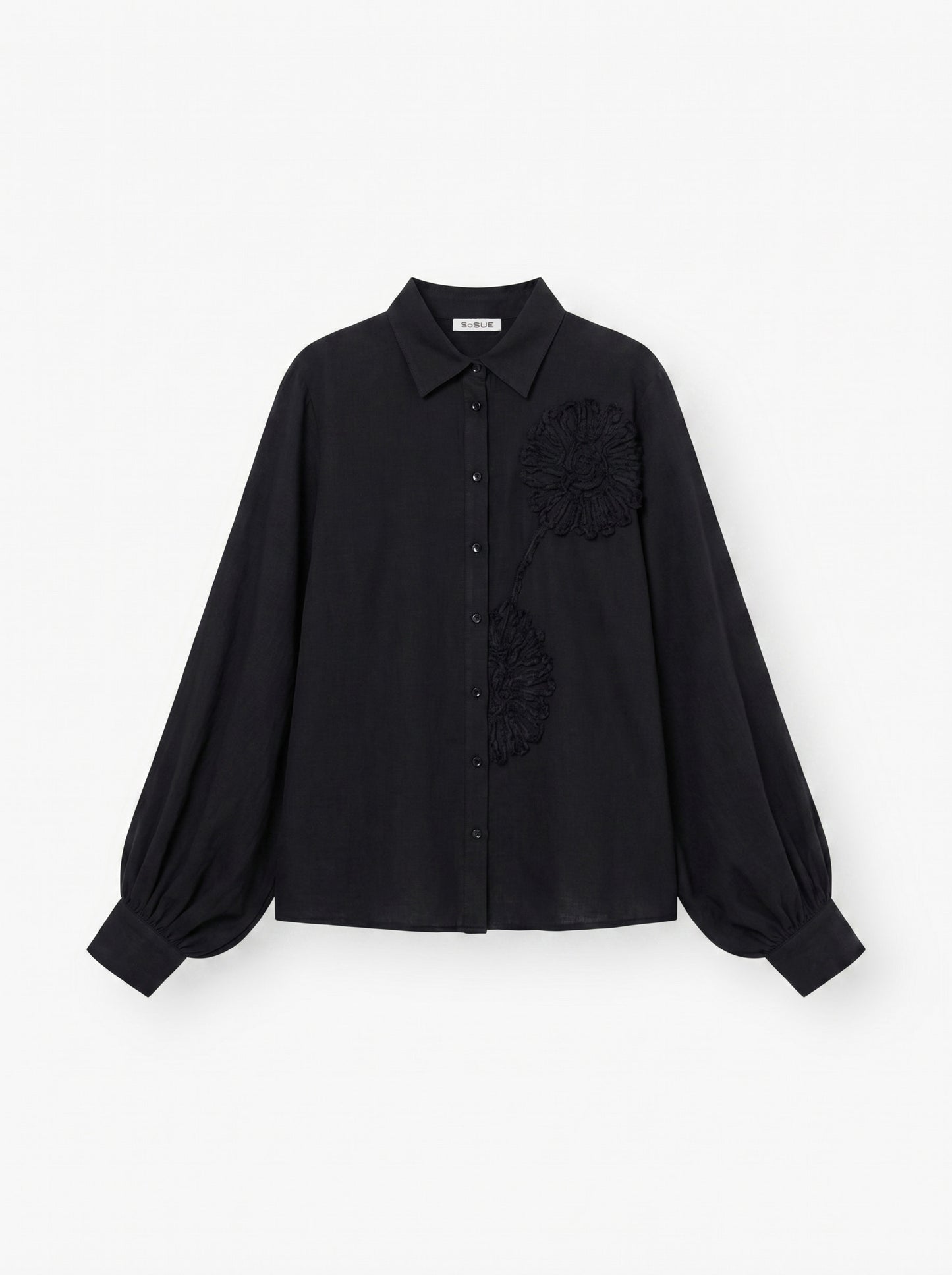Die Antonia Crafted Black ist eine schwarze, langärmelige Button-up-Bluse mit Kragen, voluminösen Ärmeln und zwei strukturierten Blumenapplikationen auf der Vorderseite.
