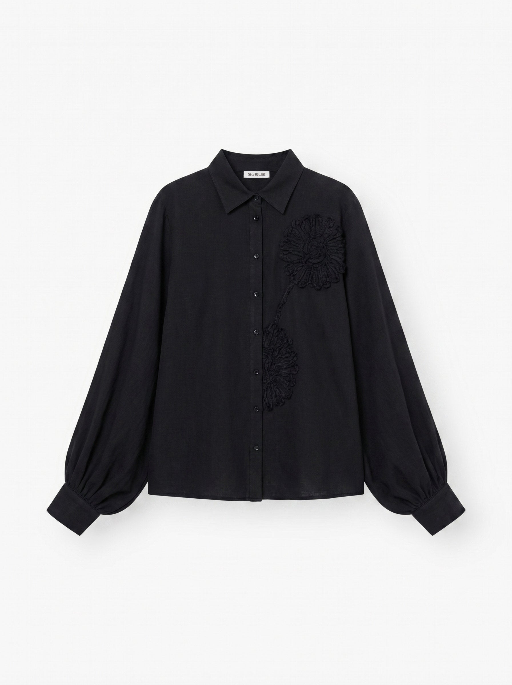 Die Antonia Crafted Black ist eine schwarze, langärmelige Button-up-Bluse mit Kragen, voluminösen Ärmeln und zwei strukturierten Blumenapplikationen auf der Vorderseite.