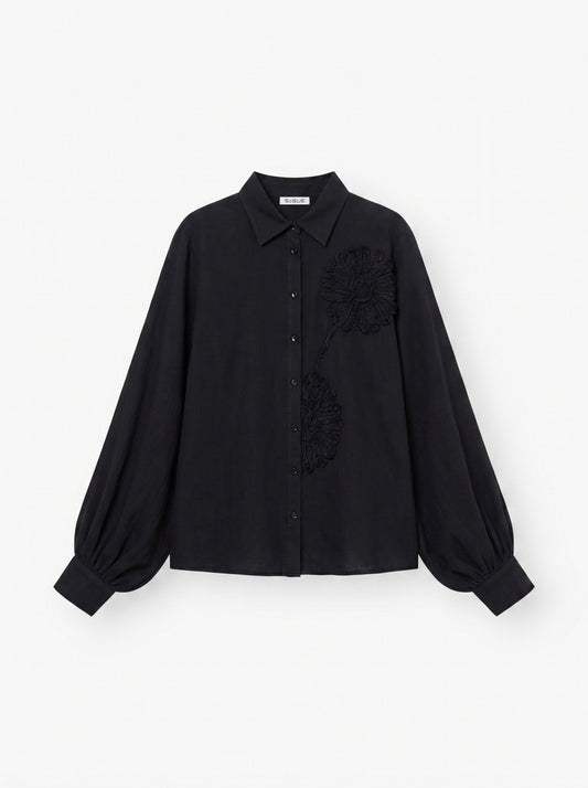 Die Antonia Crafted Black ist eine schwarze, langärmelige Button-up-Bluse mit Kragen, voluminösen Ärmeln und zwei strukturierten Blumenapplikationen auf der Vorderseite.