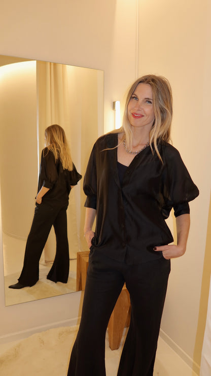 Eine Frau mit langen blonden Haaren, die ein Bluse Antonia Short Sleeve Black und eine schwarze Hose trägt, steht lächelnd mit den Händen in den Taschen da. Ihr Spiegelbild erscheint in einem hohen Spiegel hinter ihr in einem sanft beleuchteten Raum.