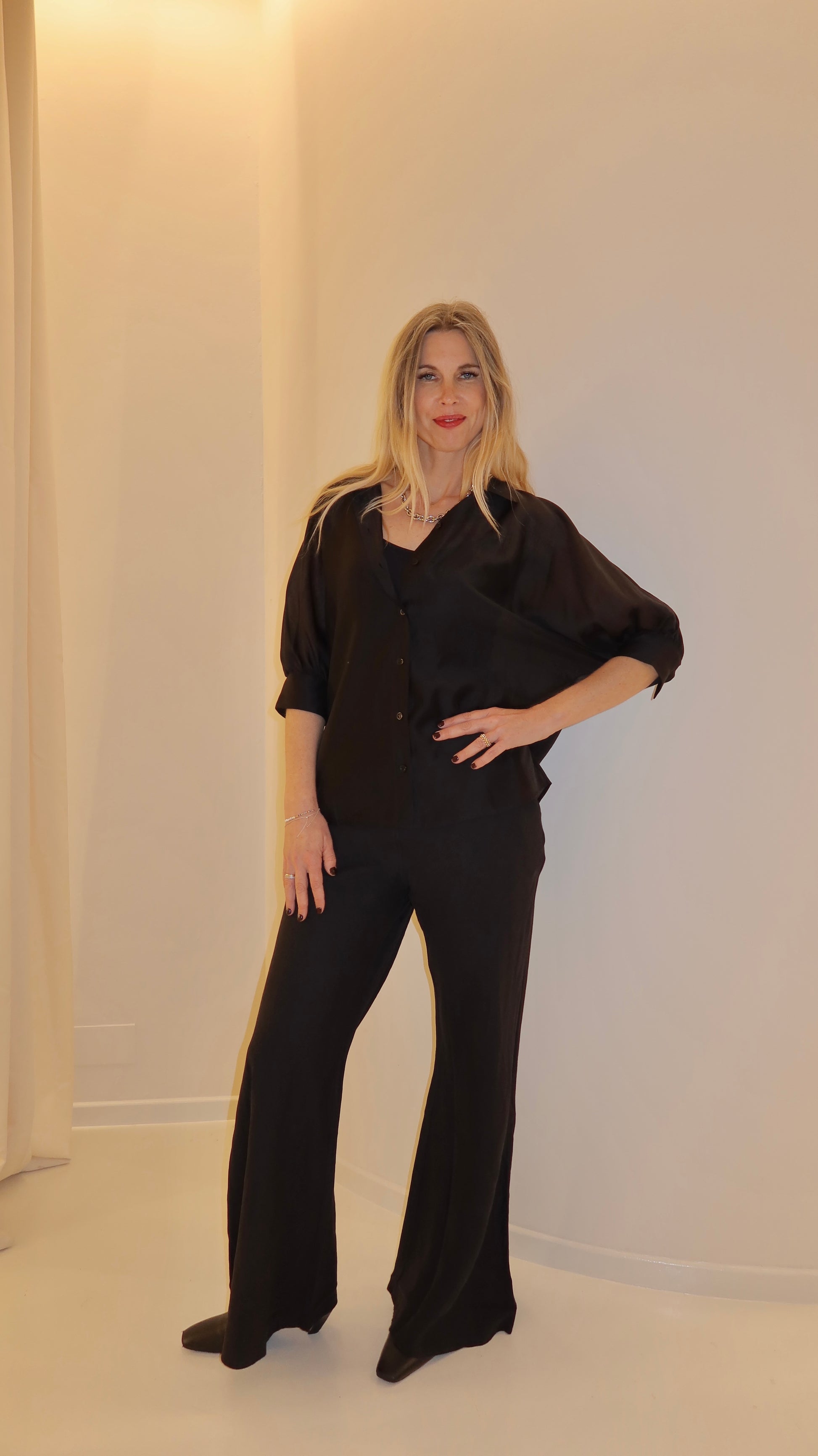 Eine Frau mit langen blonden Haaren steht vor einer hellen Wand. Sie trägt das Bluse Antonia Short Sleeve Black und eine passende Hose mit weitem Bein. Sie posiert mit einer Hand auf ihrer Hüfte und lächelt leicht.