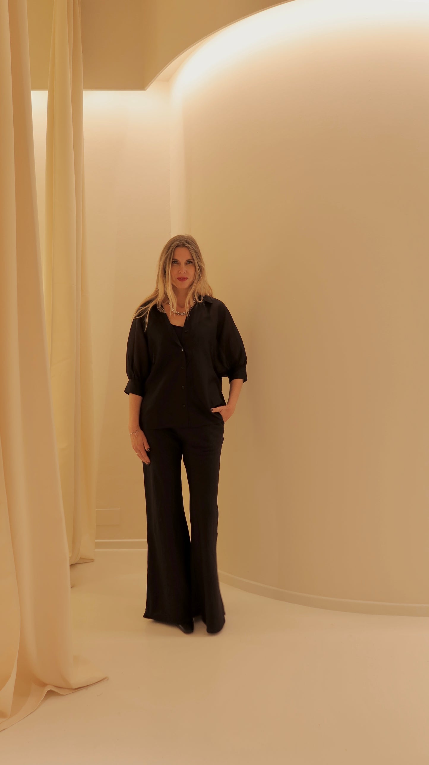 Eine Frau mit langen blonden Haaren steht in einem minimalistischen cremefarbenen Raum, trägt die Bluse Antonia Short Sleeve Black und eine schwarze Hose mit weitem Bein, eine Hand in der Tasche und einen neutralen Blick.