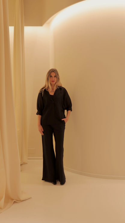 Eine Frau mit langen blonden Haaren steht in einem minimalistischen cremefarbenen Raum, trägt die Bluse Antonia Short Sleeve Black und eine schwarze Hose mit weitem Bein, eine Hand in der Tasche und einen neutralen Blick.