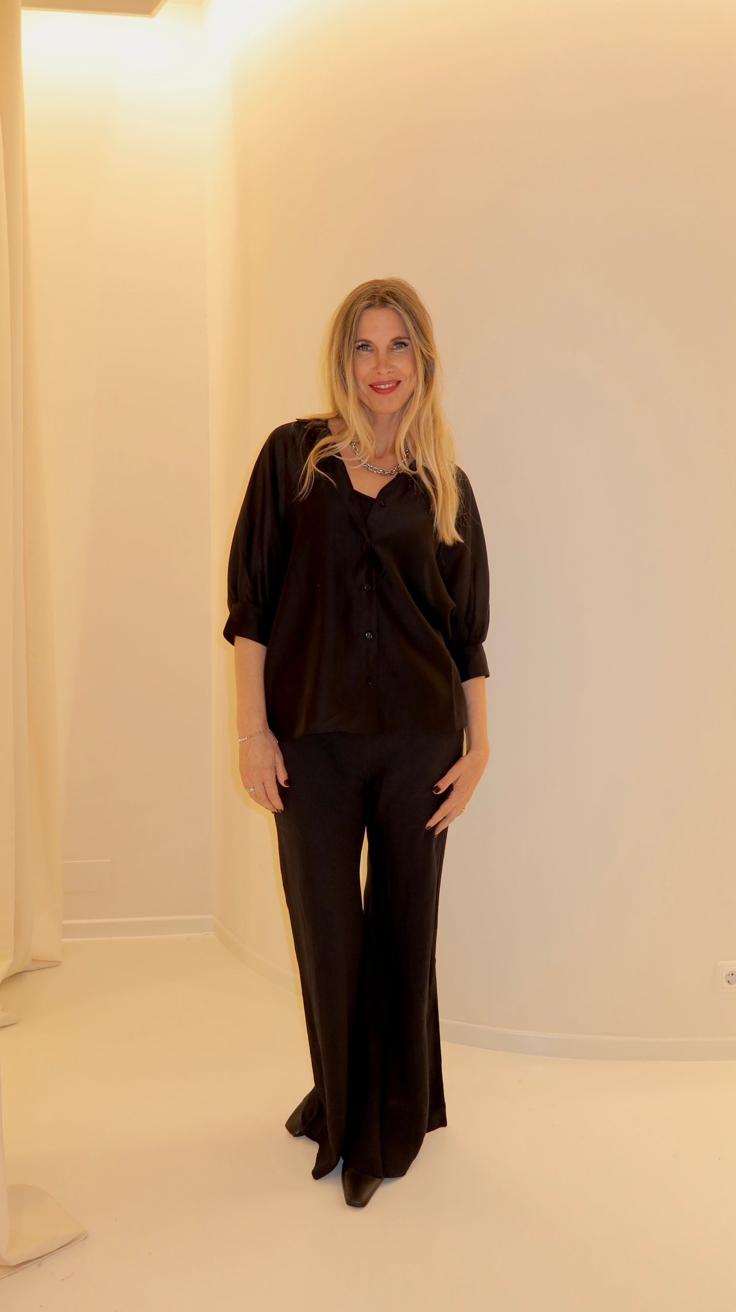 Eine lächelnde Frau mit langen blonden Haaren steht drinnen vor einer weißen Wand. Sie trägt das Bluse Antonia Short Sleeve Black im Oversized-Schnitt mit einer passenden Hose mit weitem Bein und schwarzen Schuhen. Warmes, weiches Licht erfüllt die Szene.