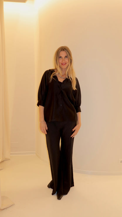 Eine lächelnde Frau mit langen blonden Haaren steht drinnen vor einer weißen Wand. Sie trägt das Bluse Antonia Short Sleeve Black im Oversized-Schnitt mit einer passenden Hose mit weitem Bein und schwarzen Schuhen. Warmes, weiches Licht erfüllt die Szene.