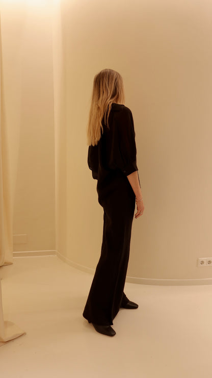 Eine Frau mit langen blonden Haaren steht in einem Innenraum mit dem Rücken zur Kamera. Sie trägt das übergroße Bluse Antonia Short Sleeve Black und eine schwarze Hose. Die minimalistische Szene ist vor einer beigen Wand sanft beleuchtet.