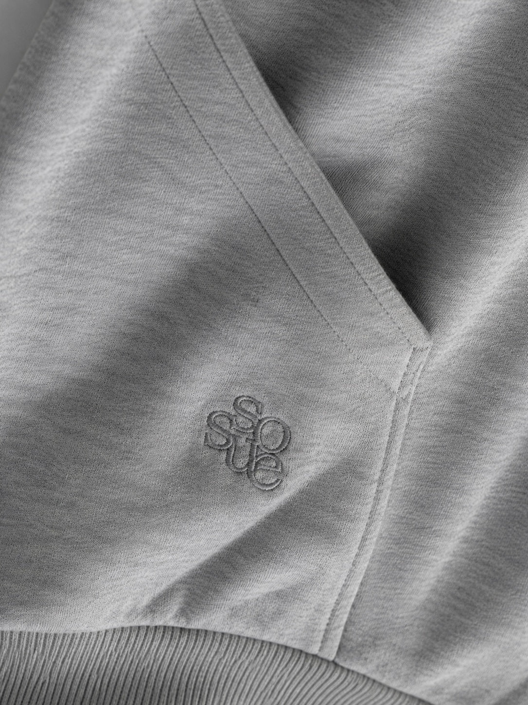 Nahaufnahme des „Grauer Oversized Hoodie“ aus hellgrauem Stoff mit einer Tasche und einer dezenten „sosue“-Stickerei in der Nähe der Naht. Dieser Oversized-Hoodie aus weichem, leicht strukturiertem Fleece wird in Portugal hergestellt.