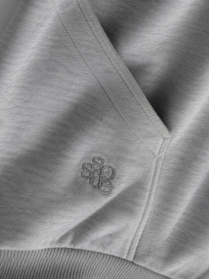 Nahaufnahme des „Grauer Oversized Hoodie“ aus hellgrauem Stoff mit einer Tasche und einer dezenten „sosue“-Stickerei in der Nähe der Naht. Dieser Oversized-Hoodie aus weichem, leicht strukturiertem Fleece wird in Portugal hergestellt.