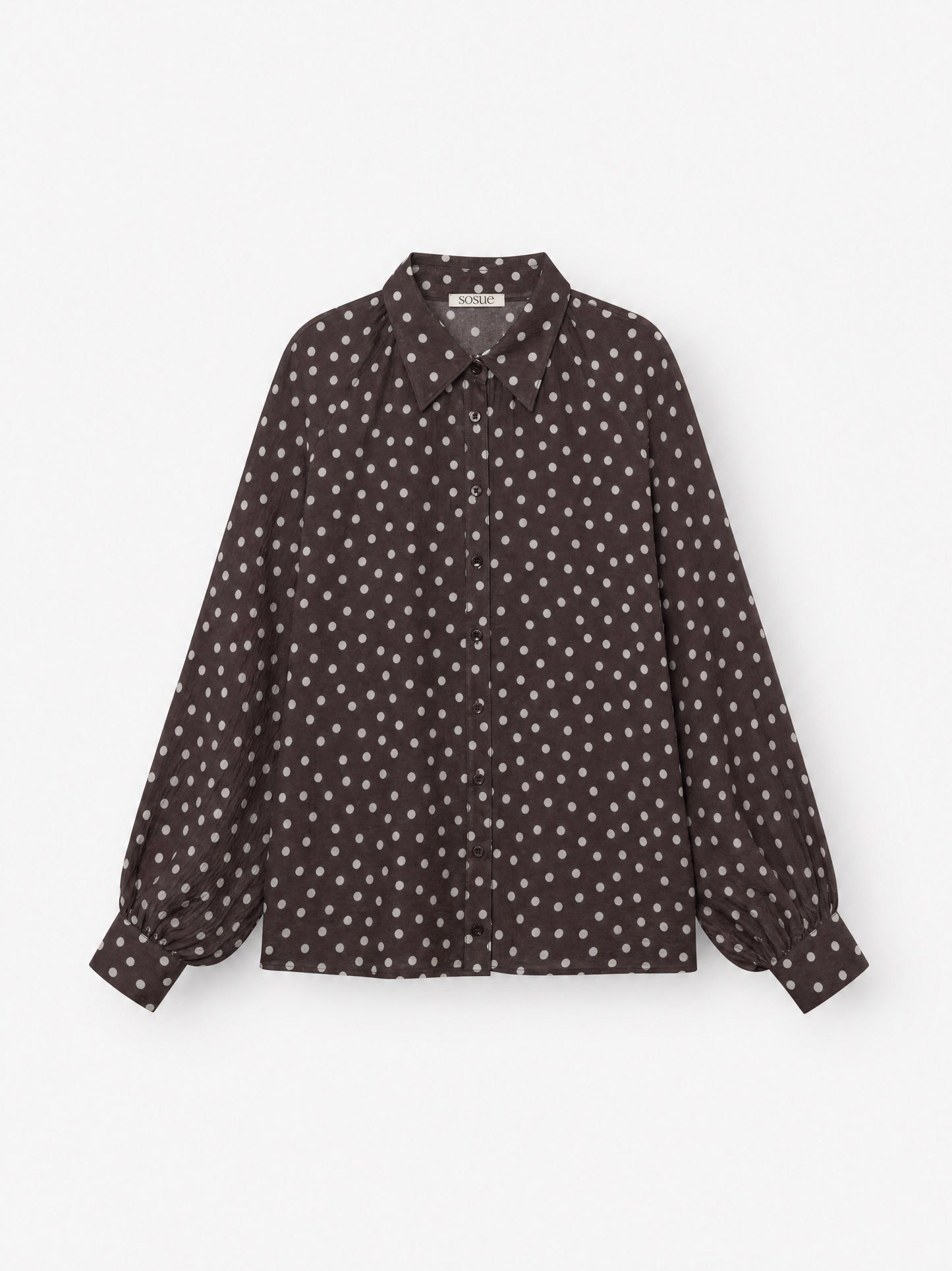 Die Bluse „Antonia Polka Dots Brown Ecru“ ist eine langärmelige Damenbluse mit Knopfleiste aus Baumwoll-Voile, die sich durch ein dunkelbraun-weißes Polka-Dot-Muster mit spitzem Kragen auszeichnet, das auf einem schlichten weißen Hintergrund abgebildet ist.