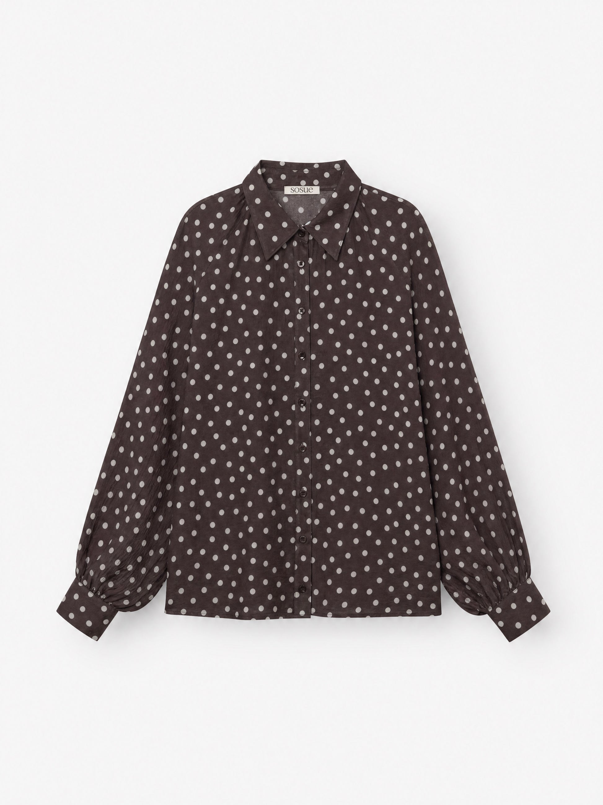 Die Bluse „Antonia Polka Dots Brown Ecru“ ist eine langärmelige Damenbluse mit Knopfleiste aus Baumwoll-Voile, die sich durch ein dunkelbraun-weißes Polka-Dot-Muster mit spitzem Kragen auszeichnet, das auf einem schlichten weißen Hintergrund abgebildet ist.