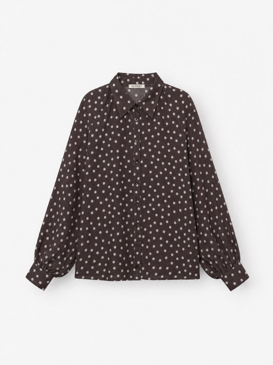 Die Bluse „Antonia Polka Dots Brown Ecru“ ist eine langärmelige Damenbluse mit Knopfleiste aus Baumwoll-Voile, die sich durch ein dunkelbraun-weißes Polka-Dot-Muster mit spitzem Kragen auszeichnet, das auf einem schlichten weißen Hintergrund abgebildet ist.