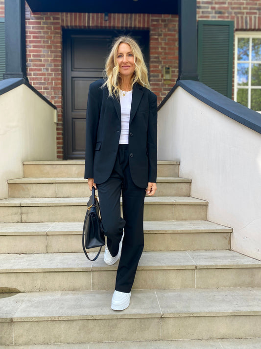 Auf einer Freitreppe trägt eine Frau mit langen blonden Haaren eine Hose von Karoline Black, die mit einem weißen T-Shirt, einem schwarzen Blazer und weißen Turnschuhen kombiniert ist. Sie hält eine schwarze Handtasche in der Hand und steht leicht lächelnd vor einem Backsteingebäude mit einer dunklen Tür.